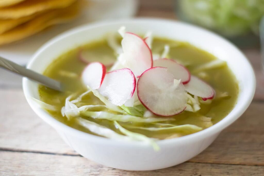 green chicken pozole recipe in a white bowl with sliced radishes and shredded cabbage.