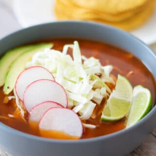 Red chicken pozole in a bowl with shredded cabbage, sliced radishes, avocado and fresh cut limes.